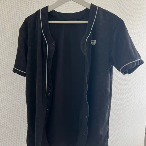 Svart kortärmad tröja från FSBN - En svart kortärmad tröja från FSBN med vita detaljer och knappar framtill. Tröjan har ett diskret mönster och vita kanter vid ärmslut och hals.