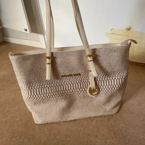 Beige handväska från Michael Kors - En elegant beige handväska från Michael Kors med ett ormskinnsmönster. Väskan har två långa handtag och en avtagbar axelrem. Den har en dragkedja och flera innerfickor för förvaring. kan ej garantera äktheten!