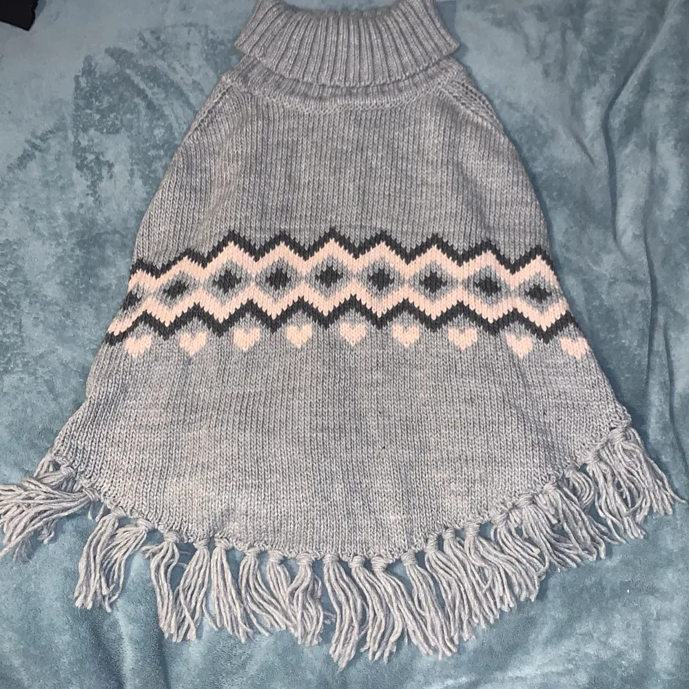 Säljer en superfin grå stickad poncho med ett mönster i rosa och svart. Den har en hög krage och fransar längs nederkanten. Perfekt för höst och vinter! 🧣. Tröjor & Koftor.