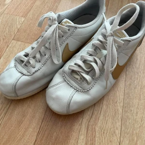 Vita Nike sneakers med guldlogga - Ett par vita Nike sneakers med guldlogga på sidan och hälen. Skorna har snörning och är tillverkade i skinn.