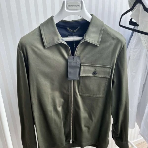 Olivgrön overshirt från J.Lindeberg - Säljer en snygg olivgrön overshirt från J.Lindeberg. Den har en dragkedja framtill och en bröstficka med knapp. Perfekt för vår och höst, och kan bäras både som en jacka eller som en skjorta. Materialet känns mjukt och bekvämt, och passformen är normal. Perfekt för en stilren och avslappnad look!