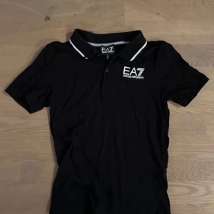 Svart pikétröja från Emporio Armani - Säljer en snygg svart pikétröja från Emporio Armani. Tröjan har en klassisk krage med vita detaljer och knappar framtill. Perfekt för både vardag och fest!