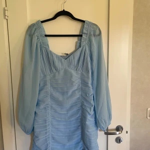 Ljusblå klänning med långa ärmar - Säljer en superfin ljusblå klänning med långa ärmar. Klänningen har en v-ringad design och är gjord av ett lätt och luftigt material, perfekt för vår och sommar. Endast provad annars helt oanvänd!