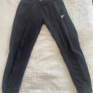 Svarta mjukisbyxor från Nike - Svarta mjukisbyxor från Nike med broderad vit logga på vänster ben. Byxorna har resår och snörning i midjan samt muddar vid bensluten. Tillverkade i mjukt bomullsmaterial, perfekta för chill eller träning. Noppriga på insidan och färgen på  annars i fint skick metallen vid snörningen har börjat gå bort🤍