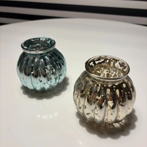 Glaslyktor - Bilderna visar två dekorativa glaslyktor i rundad form, en i blått och en i silverfärgat glas med antik finish. De har ett vackert, reflekterande mönster och passar perfekt som värmeljushållare eller dekoration. Inga synliga sprickor eller större skador, endast naturligt slitage.