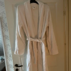 Vit morgonrock i frotté med bälte - Mysig vit morgonrock i mjuk frotté med långa ärmar och knytskärp i midjan. Perfekt att slänga på sig efter duschen eller när du vill chilla hemma. Klassisk modell med skön känsla och enkel design.