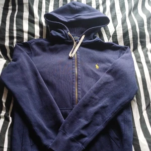 Marinblå hoodie från Polo Ralph Lauren - Snygg marinblå hoodie från Polo Ralph Lauren med dragkedja och huva. Klassisk gul logga på bröstet och vita snören i luvan. Perfekt för dig som gillar en stilren och sportig look. Tillverkad i mjuk bomull för extra komfort.
