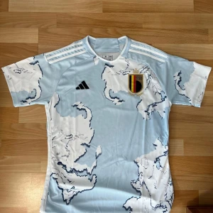 Belgien bortatröja Adidas M - Snygg Belgien bortatröja från Adidas i slim fit. Tröjan är ljusblå och vit med ett unikt abstrakt mönster, klassiska Adidas-ränder på axlarna och Belgiska landslagets emblem på bröstet. Tillverkad i lätt och ventilerande polyester, perfekt för match eller träning. Helt ny!