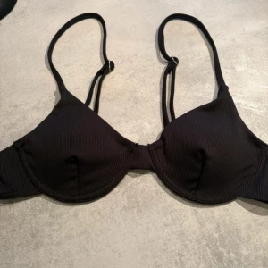 Bikiniöverdel - Snygg svart bikiniöverdel med ribbad struktur och vadderade kupor. Justerbara axelband och metallspänne bak för perfekt passform. Enkel och stilren design som passar till alla badtillfällen.