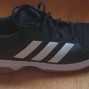 Svarta Adidas idrottsskor strl 44 - Säljer ett par svarta Adidas innomhus-idrottsskor Skorna har snörning, mesh- och syntetmaterial samt gummisula med non-marking-funktion. Sparsamt använda. Storlek 44.