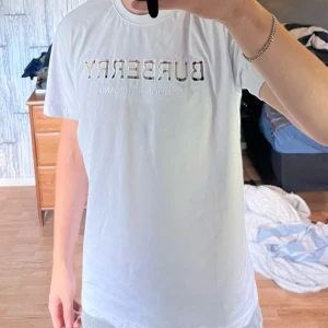 Vit Burberry t-shirt - Säljer en fräsch vit Burberry t-shirt i 100% bomull. T-shirten har korta ärmar och rund hals. På bröstet finns en broderad Burberry-logga med klassiskt rutmönster i bokstäverna och texten 'London England' under. Perfekt för dig som gillar stilrena designer med lyxig touch. Använd 1 gång