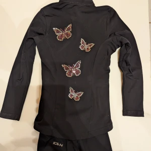 Svart träningsjacka och  byxor ICE H med fjärilar - Svart träningsjacka från ICE H med glittrande fjärilar i strass på ryggen och ena ärmen. Jackan har dragkedja framtill, lång ärm och liten svensk flagga i strass på bröstet. Snygg och sportig passform i stretchigt syntetmaterial.storlek 120-130