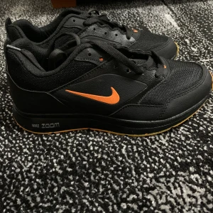 Nike Zoom - ’’INTE OG’’.                                                        Svarta Nike Zoom sneakers med orange swoosh och detaljer. Ovandel i mesh och syntet, snörning och platt sula. Sulan är orange undertill och skon har tydliga sportiga vibbar. Perfekta för dig som gillar en clean men ändå färgstark look.
