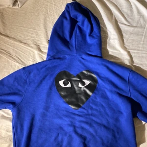 Blå Comme des Garçons PLAY hoodie L - Blå hoodie från Comme des Garçons PLAY med dragkedja och huva. Ikoniskt svart hjärta med ögon på bröstet och stort tryck på ryggen. Mjuk bomullskvalitet och praktiska fickor framtill. Perfekt för dig som vill sticka ut med streetstyle.