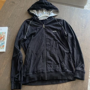 Svart hoodie med dragkedja från BOSS  - Svart tunnare hoodie från BOSS Homewear med vit piping längs axlarna och grått foder i huvan. Tröjan har dragkedja framtill, två fickor och BOSS-logga på bröstet. 