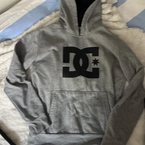 Grå DC hoodie med svart logga - Säljer en grå hoodie från DC med stor svart logga på bröstet. Tröjan har huva, magficka och långa ärmar. Perfekt för chill dagar och streetwear-stil. Materialet är mjuk bomull och polyesterblandning som känns skönt mot huden. Har ett litet hål(se bild)