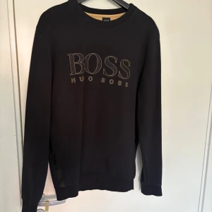 Svart sweatshirt från Hugo Boss - Svart sweatshirt från Hugo Boss med guldfärgad logga framtill. Klassisk rund halsringning och långa ärmar. Tröjan har ribbade muddar och är tillverkad i mjuk bomullsmix för en skön känsla. Perfekt för en clean och stilren look.
