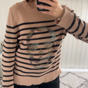 Randig Zadig & Voltaire tröja med döskalle - Beige och svart randig stickad tröja från Zadig & Voltaire med glittrig döskalle framtill. Tröjan har rund hals, långa ärmar och snygga knappar på axeln. Perfekt för dig som gillar edgy detaljer och cool stil.