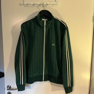 Grön ziptröja från Lacoste - Grön track jacket från Lacoste med diskret mönster och vita ränder längs ärmarna. Jackan har dragkedja framtill, ståkrage och ribbade muddar. Klassisk Lacoste-logga på bröstet. Perfekt för en sportig och stilren look.