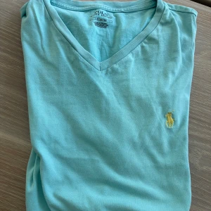 Turkos v-ringad t-shirt från Polo Ralph Lauren - Turkos t-shirt från Polo Ralph Lauren med klassisk v-ringning och gul broderad logga på bröstet. Tillverkad i mjuk bomull, perfekt för dig som gillar stilrena och fräscha plagg. Passformen är normal och t-shirten har korta ärmar.