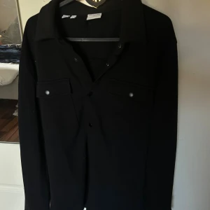Svart overshirt från selected home  - Svart overshirt från selected home med klassisk krage, två bröstfickor med lock och tryckknappar. Långärmad modell i ett mjukt material, perfekt att slänga över en t-shirt. Enkel och stilren design som funkar till det mesta. Använd max 1 gång 