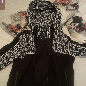 Svart vindjacka  - Svart vindjacka med vita grafiska mönster längs ärmar och axlar. Jackan har dragkedja framtill och huva.  Tar emot Byten 