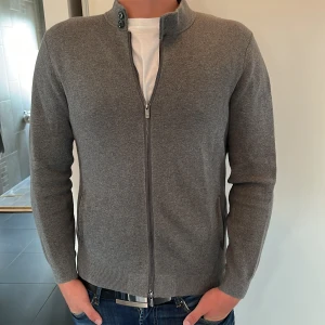 Grå kofta från Massimo Dutti - Snygg grå kofta från Massimo Dutti i extra fin bomull. Modellen har hel dragkedja framtill, ståkrage med två knappar och ribbade muddar. Perfekt till jeans och t-shirt för en clean och stilren look.
