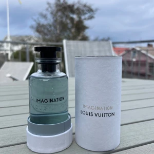 Louis Vuitton Imagination parfym - Lyxig parfym från Louis Vuitton, Imagination, med stilren glasflaska och svart lock med LV-logga. Parfymen har en ljusblå ton och kommer i en vit cylinderformad förpackning med silverdetaljer och präglad logotyp. Perfekt för dig som gillar exklusiva dofter.