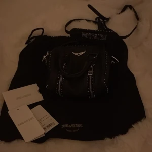 Svart handväska från Zadig & Voltaire - Cool svart handväska i skinn från Zadig & Voltaire med silvriga metalldetaljer, dubbla handtag och avtagbar axelrem. Väskan har dekorativa dragkedjor framtill, nitar längs kanterna och ikonisk vinge i metall. Perfekt storlek för det viktigaste. Köpt av en annan här på plick för 1850 