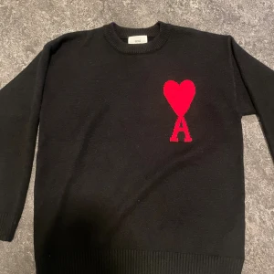Svart stickad tröja från Ami Paris - Svart stickad tröja från Ami Paris med rund hals och lång ärm. På bröstet finns ett stort rött hjärta med bokstaven A, vilket ger en ikonisk och trendig look. Perfekt för dig som gillar stilrena plagg med en twist.