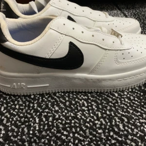 Nike Air Force 1 vit/svart strl 38 - ’’INTE OG’’.                                                        Säljer ett par klassiska Nike Air Force 1 i vitt med svarta detaljer och swoosh. Skorna har perforerad tå, snörning med metallbricka och vit platt sula. Tillverkade i skinn och syntet, med svart Nike Air-logga på hälen. Perfekt för dig som gillar streetstyle.