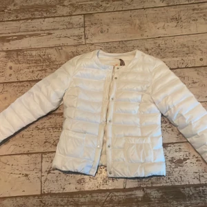 Vit pufferjacka från Holly & Whyte - Säljer en stilren vit pufferjacka från Holly & Whyte by Lindex. Jackan har rund halsringning, tryckknappar framtill och är quiltad för extra värme. Perfekt för kyliga dagar och lätt att matcha med olika outfits.