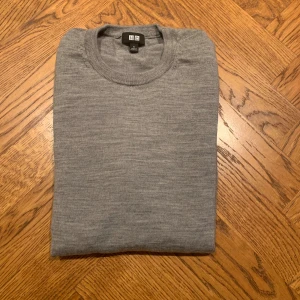 Grå merinoulltröja från Uniqlo - Säljer en stilren grå tröja från Uniqlo i mjuk merinoull. Klassisk rund halsringning och långärmad modell, perfekt för lager-på-lager under kyliga dagar. Enkel och clean look som funkar till det mesta. Den har ett litet hål i högra armen men inget som förändrar mycket annars.