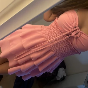 Rosa volangklänning med snörning - Supersöt rosa kortklänning med flera volanger och smockad midja. Axelbandslös modell med snörning framtill som ger en snygg detalj. Perfekt för sommarens alla tillfällen och ger en riktigt gullig vibe. (Går att använda från storlek XS till M