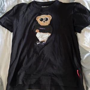 Svart t-shirt med björn från Members Only - Svart t-shirt från Members Only med broderad björn på bröstet. Klassisk passform och rund halsringning. Tillverkad i mjuk bomull som känns skön mot huden. Perfekt för dig som gillar streetstyle och coola detaljer.