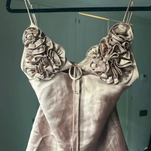 Champagnefärgad kortklänning med volang - Supersöt kortklänning i champagnefärgad satin med smala axelband och stora volangdetaljer vid bysten. Klänningen har en snygg omlottkänsla framtill med dekorativ knytning och en lätt glansig finish. Perfekt för dig som vill sticka ut med en unik och trendig look.