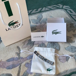Silverfärgat armband från Lacoste - Stilrent armband från Lacoste i silverfärg med länkdesign och ingraverade detaljer. Armbandet har den klassiska krokodil-loggan på en av länkarna och levereras i originalpåse och ask. Perfekt accessoar för dig som gillar sportig och clean stil. Längden på armbandet är 20-21cm.  