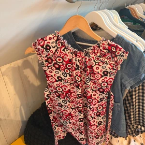 Mönstrad topp från Roco - Säljer en topp från Roco med ett coolt cirkelmönster i rött, svart och vitt. Klänningen har elastisk halsringning med volangdetaljer och knyt i midjan. Materialet är 65% bomull och 35% polyester, vilket gör den både mjuk och lätt. Perfekt för dig som gillar färg och mönster!