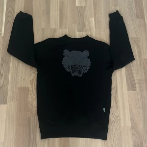 Svart Kenzo sweatshirt med tigertryck - Svart sweatshirt från Kenzo med ikoniskt tigertryck i mörkgrått framtill. Tröjan har rund halsringning och långa ärmar. Perfekt för dig som gillar streetstyle och vill ha något som sticker ut men ändå är stilrent.