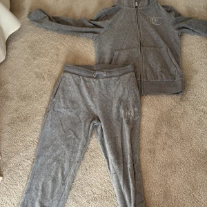 Grå hoodie från Juicy Couture - Mysig grå hoodie från Juicy Couture i mjuk velour med dragkedja framtill och huva. Tröjan har långa ärmar, fickor och glittrande JC-logga i strass på bröstet. Perfekt för chill dagar eller när du vill ha en avslappnad look.