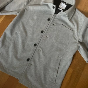 Grå overshirt från Lindbergh - Super snygg overshirt från Lindbergh i regular fit. Jackan har klassisk krage, svarta knappar framtill, bröstficka och två sidofickor. Tillverkad i mjukt bomullsmaterial med en clean look som passar perfekt till lager-på-lager.