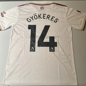 Arsenal Gyökeres 14 bortatröja Adidas - Snygg Arsenal bortatröja med Gyökeres och nummer 14 på ryggen. Tröjan är vit med vinröda detaljer, broderat klubbmärke och Adidas-logga. Tillverkad i lätt och ventilerande polyester med diskret geometriskt mönster. Klassisk krage och korta ärmar. Replika, endast handtvätt gäller!