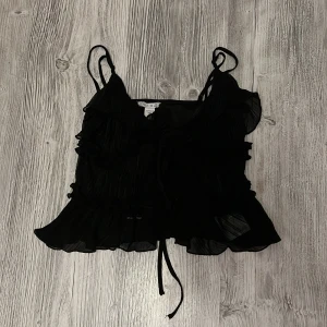 Svart volangig linnetopp från Amisu - Supersöt svart linnetopp från Amisu med tunna axelband, volangdetaljer och knyt framtill. Toppen är lätt transparent och har en plisserad struktur som ger en cool look. Perfekt att styla med jeans eller kjol för en trendig vibe.