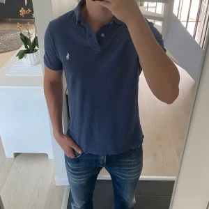 Blå pikétröja från Polo Ralph Lauren - Säljer en klassisk blå pikétröja från Polo Ralph Lauren med vit broderad logga på bröstet. Tröjan har korta ärmar, krage och knappar vid halsen. Tillverkad i mjuk bomull som är skön att bära. Perfekt för en stilren och avslappnad look.