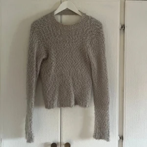 Beige omlottstickad tröja  från Nelly - Mysig beige stickad tröja från Nelly med omlottmodell och lång ärm. Tröjan har en fluffig struktur och är tillverkad i en mix av nylon, polyester och akryl. 