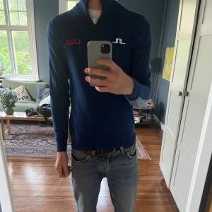 Blå half zip polotröja J.Lindeberg USA - Snygg marinblå polotröja från J.Lindeberg med half zip och broderad USA-text på bröstet och ryggen. Tröjan har långa ärmar med coola randiga detaljer och är tillverkad i mjuk bomull. Perfekt för dig som gillar sportig och stilren look. Passar även S. 