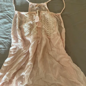 Ljusrosa topp med spets - Supersöt ljusrosa topp med virkade spetsdetaljer framtill och flätade axelband. Toppen är ärmlös och har en luftig, flowy passform som passar perfekt till varma dagar. Dekorativ knytning med tofsar framtill ger en extra boho-touch.