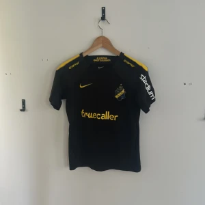 AIK fotbollströja Nike Dri-Fit svart L - AIK fotbollströja från Nike i svart med gula detaljer och klubbmärke på bröstet. Tröjan har korta ärmar, Dri-Fit-material för bra andningsförmåga och tryck från sponsorer som Truecaller och SVEA. Perfekt för dig som vill visa ditt AIK-stöd.