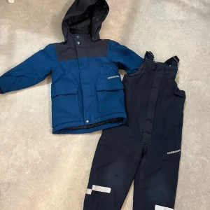 Blå Didriksons vinterjacka med huva - Säljer en blå och mörkblå vinterjacka från Didriksons med huva och stora fickor framtill. Jackan är 100% vattentät och har dragkedja samt knappar. Perfekt för kalla dagar och har en sportig look. Märket syns tydligt på bröstet.