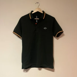 Olivgrön Replay pikétröja med orange detaljer - Olivgrön pikétröja från Replay med orange detaljer på krage och ärmslut. Klassisk pikékrage med Replay-logga i orange på baksidan av kragen. Liten logotyp på bröstet. Skön passform och tillverkad i mjuk bomull.